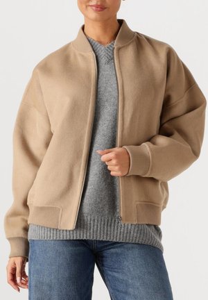 Beige Bomberjacke mit Reißverschluss vorne, gerippten Bündchen und Saum. Getragen über einem grauen Pullover mit strukturierter Musterung und normaler Passform.
