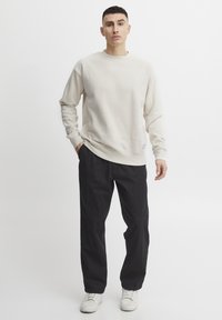 Sudadera beige de manga larga y cuello redondo, combinada con pantalones negros de corte holgado y zapatillas blancas. Detalle de etiqueta sutil en la sudadera.
