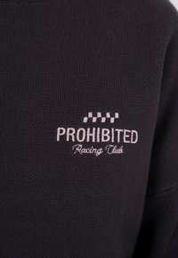 Sweat-shirt noir tricoté avec un logo brodé rose, des accents à damier et l'inscription "PROHIBITED Racing Club."