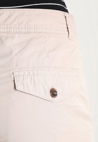 Shorts en tissu rose clair avec une poche arrière, fermée par un bouton marron. Détail de couture visible le long des bordures. Texture douce.