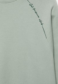Sweat-shirt vert clair avec broderie cursive vert foncé le long de la couture de l'épaule indiquant "Club boutique club".