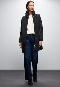 Schwarzer Mantel mit hohem Kragen, Druckknopfverschluss vorne, getragen über einem cremefarbenen Kabelstrickpullover und dunklen weit geschnittenen Jeans. Schwarze Schuhe.