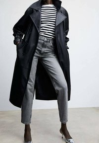 Personne portant un trench-coat en cuir noir, une chemise rayée noir et blanc, un jean gris taille haute et des chaussures à talons argentées, debout les mains dans les poches.