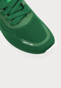 Zöld sportcipő hálós felsőrésszel, lapos fűzőkkel és fehér gumitalppal. Kiemelkedő részlet: "1890 óta" felirat az oldalon.