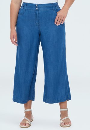 Donna che indossa pantaloni in denim blu cropped a vita media e gamba larga, con chiusura a doppio bottone e sandali beige con cinturino alla caviglia.