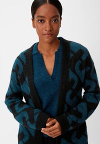 Strickjacke in Petrol und Schwarz mit einem auffälligen geometrischen Muster, ausgestattet mit einem V-Ausschnitt und langen Ärmeln, aus weichem, flauschigem Material.