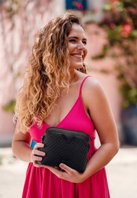 Black quilted bag with a structured design, featuring a V-shaped accent, held in hands. Model wears a pink dress with thin straps.