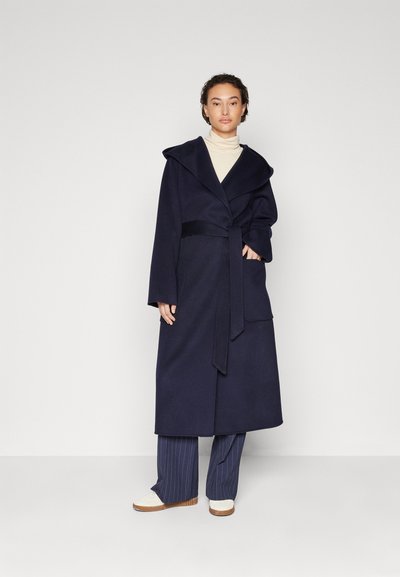 Cappotto di lana navy con una grande cappuccio, cintura autoinserita e tasche laterali, indossato sopra un leggero dolcevita e pantaloni a righe.