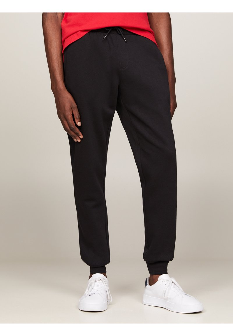 Svarta sweatpants i mjukt tyg med dragsko i midjan, ribbade muddar och sidofickor, kombinerat med vita skor och en röd skjorta.