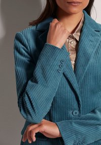 Teal corduroy blazer met een geknipt kraag, geribbelde textuur en drie blauwe knopen op de manchetten. Gelaagd over een beige satijnen shirt.