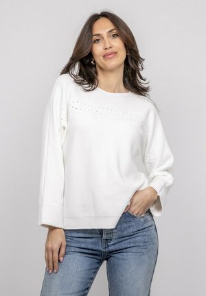 Femme aux cheveux bruns mi-longs portant un pull blanc à manches longues avec de petits détails, un jean bleu, posant devant un fond uni.