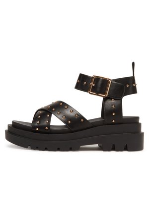 Black leather sandal with studded accents, a chunky platform, and an ankle strap featuring a gold buckle. Textured sole for grip.
