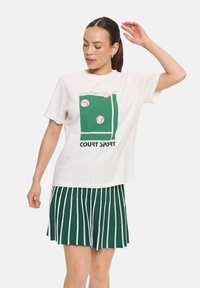 T-shirt en coton blanc avec un design graphique vert représentant des balles de tennis et du texte, associé à une jupe plissée rayée verte et blanche.