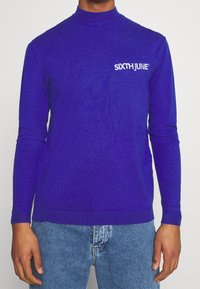 Lila långärmad tröja med mock neck, stickad textur och vit "SIXTH JUNE"-logotyp på bröstet. Matchad med blå denimjeans.