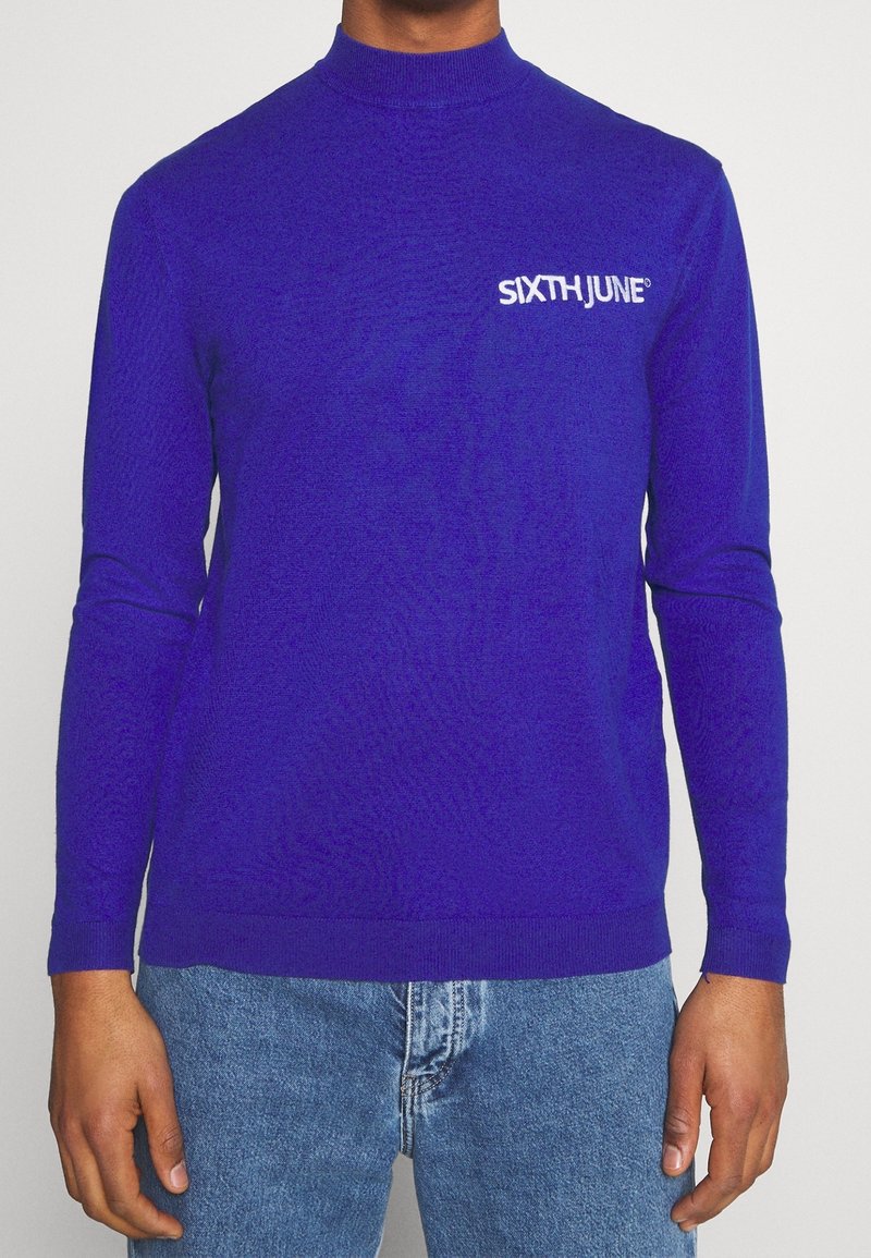 Lila långärmad tröja med mock neck, stickad textur och vit "SIXTH JUNE"-logotyp på bröstet. Matchad med blå denimjeans.