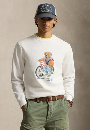 Młody mężczyzna w kremowej bluzie z nadrukiem misia i roweru, zielonych spodniach, brązowym pasku, niebieskim zegarku i granatowej czapce Ralph Lauren.