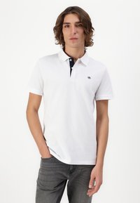 Tricou polo alb cu guler albastru închis și trei butoane, având un logo mic pe piept. Purtați cu blugi gri închis.
