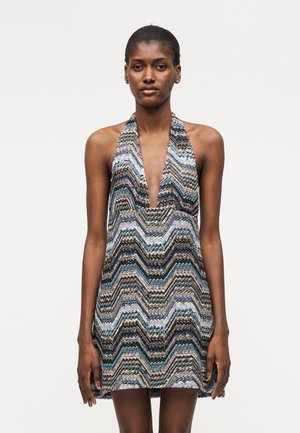 Γυναίκα που φορά φόρεμα χωρίς μανίκια με βαθύ V-λαιμόκοψη και πολύχρωμο ζιγκ ζαγκ μοτίβο σε αποχρώσεις μπλε, μαύρου, μπεζ και λευκού.