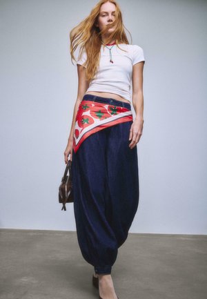 Mladá žena kráča, oblečená v bielom krátkom tričku, tmavých voľných nohaviciach, červenom kvetinovom šatke previazanom v páse a nesie malú hnedú kabelku.