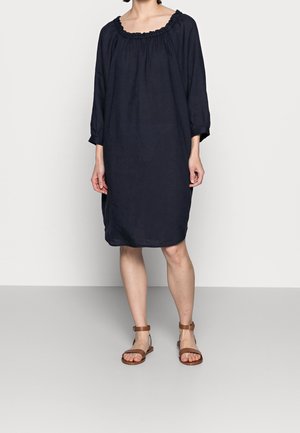 Kvinde iført en løs, knælængde navy kjole med trekvartlange ærmer og brune sandaler med ankelrem, stående mod en ensfarvet baggrund.