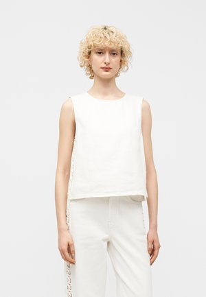 Femme aux cheveux blonds bouclés, debout face à l'avant, vêtue d'un haut blanc sans manches et d'un pantalon blanc avec des côtés à lacets.