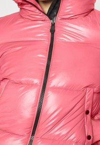 Chaqueta acolchada rosa con un cuello alto, textura brillante, cremallera frontal en negro y un bolsillo en el pecho con un botón decorativo.