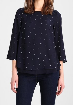 Frau trägt eine marineblaue Langarmbluse mit kleinen weißen Blumenmustern und dunkelblaue Jeans vor einem schlichten Hintergrund.