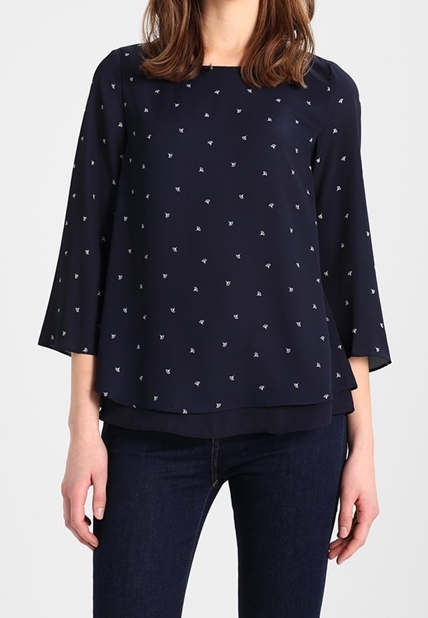 Femme portant un chemisier bleu marine à manches longues avec de petits imprimés floraux blancs et un jean bleu foncé sur un fond uni.