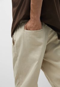 Person wearing beige pants with right hand inside front pocket, and a brown shirt hanging loosely over the waistband.