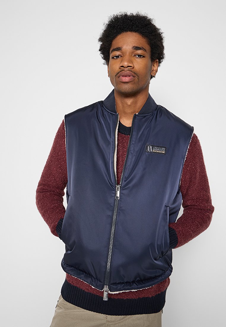 Giovane con le mani in tasca indossa un gilet blu navy con zip sopra un maglione bordeaux su uno sfondo uniforme.