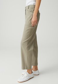 Persona con pantaloni larghi beige e sneakers bianche, in piedi di profilo contro uno sfondo chiaro uniforme.