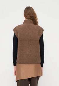 Καφέ φλις γιλέκο με ψηλό γιακά και χωρίς μανίκια, με υφή επιφάνειας, φορεμένο πάνω από μια μακρυμάνικη μαύρη μπλούζα.