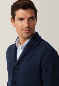 Cardigan a maglione blu navy con colletto a scialle, caratterizzato da un motivo testurizzato e tre bottoni neri, indossato sopra una camicia a righe chiara.