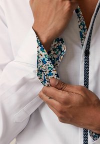Weißes Hemd aus strukturiertem Stoff, Blumenmuster an den Manschetten in Blau und Grün sowie eine kontrastierende marineblaue Knopfleiste.
