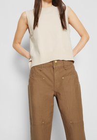 Beige mouwloos topje met een ronde hals, gecombineerd met bruine, loszittende broek met goudkleurige knoopaccenten en voorkruipzakken.