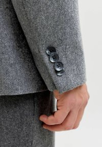 Rękaw szarej wełnianej marynarki z trzema czarnymi guzikami i kontrastującymi przeszyciami. Widoczna miękka faktura i dopasowany krój.
