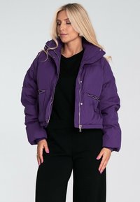 Fioletowa kurtka puchowa o skróconym kroju, błyszcząca, z zamykanymi kieszeniami i kapturem. Posiada ściągacze na mankietach oraz zamek błyskawiczny z przodu.