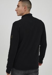 Schwarze Zip-Up-Sweatjacke mit hohem Kragen, gerippten Bündchen und kleinem gelben Logo am linken Ärmel. Aus weichem, strukturiertem Stoff gefertigt.