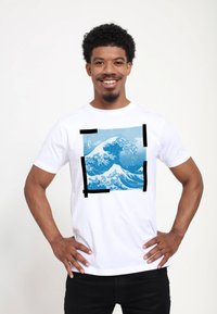 Hvid bomulds T-shirt med et blåt bølgemotiv inden for en sort rektangulær kant. Korte ærmer, rund halsudskæring. Glat tekstur.