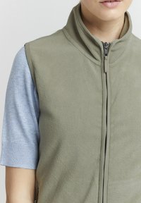 Fleece prsluk u maslinastoj zelenoj boji s visokim ovratnikom, prednjim patentnim zatvaračem i teksturiranom površinom. Nošen preko svijetloplavog majice s kratkim rukavima.