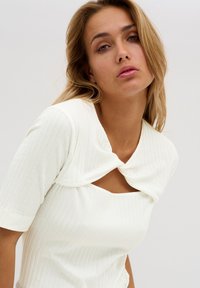 White ribbed top with a round neckline and keyhole detail, featuring short sleeves and a knotted accent at the collar. Soft texture.