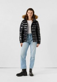 Giacca puffy nera in materiale lucido, con cappuccio foderato in pelliccia e design trapuntato verticale. Abbinata a jeans azzurri chiari e stivali neri.