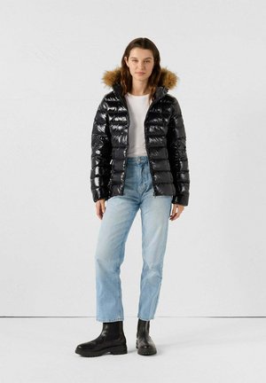 Chaqueta acolchada negra con material brillante, capucha forrada de piel y diseño de acolchado vertical. Combinada con jeans de color azul claro y botas negras.