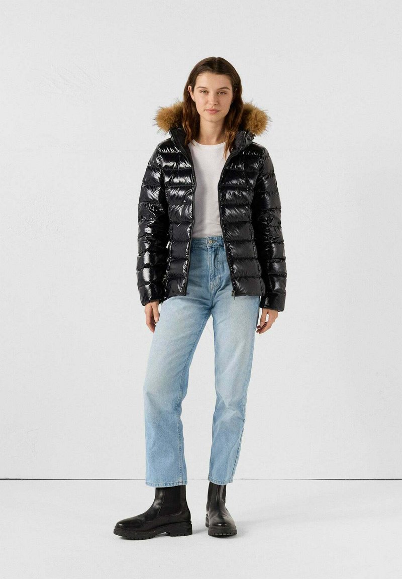 Giacca puffy nera in materiale lucido, con cappuccio foderato in pelliccia e design trapuntato verticale. Abbinata a jeans azzurri chiari e stivali neri.