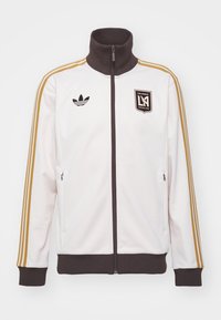 Bílá zipová bunda s hnědým límcem a manžetami. Obsahuje tři zlaté pruhy na rukávech, černé logo Adidas a emblém Los Angeles FC.