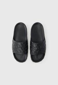 Schwarze Gummi-Slides mit einem erhabenen kreisförmigen Muster auf den Riemen und einer flachen Sohle. Der Markenname „COACH“ ist auf dem Riemen sichtbar.