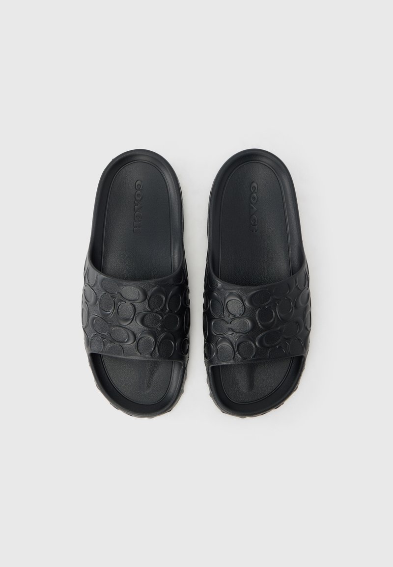 Schwarze Gummi-Slides mit einem erhabenen kreisförmigen Muster auf den Riemen und einer flachen Sohle. Der Markenname „COACH“ ist auf dem Riemen sichtbar.