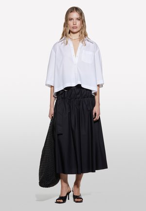 Chemisier blanc coupé avec une poche, associé à une jupe longue noire avec une taille froncée, des sandales noires à talons, et un sac en filet.