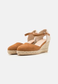 Zapatos de cuña de ante marrón con suela de yute trenzado, correa en el tobillo y un panel de tela texturizada beige en la parte trasera. Diseño de punta redondeada.