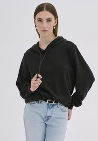 Fekete kapucnis pulcsi laza fazonnal, zsinóros kapucnival és hosszú ujjakkal, világoskék farmerrel párosítva. Simára szőtt anyag és letisztult design.